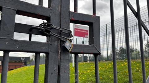 Some large black metal gates locked by a huge padlock. On the other side of the gates are a grassy bank and a pink sign