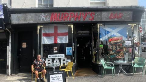 A man sits on a chair out the front of a shop. The sign at the front reads Murphy's Bar. A large England flag can be seen hanging in the window.
