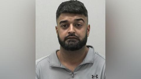 Mugshot of Ali. He has short, neat thick black hair and a black beard and is wearing a grey zip-up polo shirt.