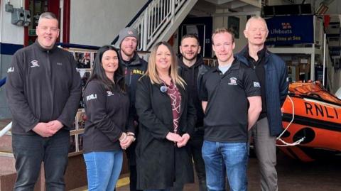 Seven people, five men and one woman stand in a lifeboat shed looking at the camera. A small lifeboat is in the background