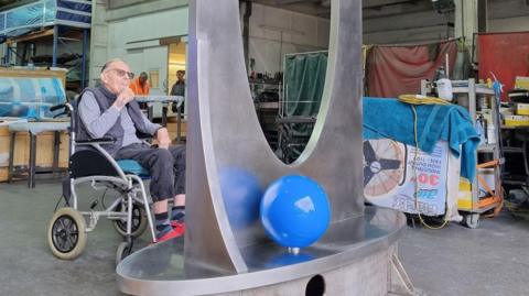An elderly man sits in a wheelchair next to the large steel structure in a busy warehouse.