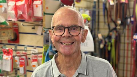Image of an older man wearing a grey polo top and black reading glasses. He is smiling at the camera. In the background can be seen racks of dog leads, collars and electrical sockets and plugs.