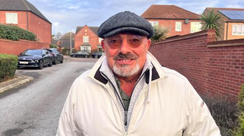 Tony stands in front of some parked cars in a residential street close to Castle Hill Hospital wearing a white coat, flat cap and glasses.