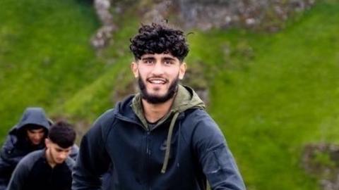 Mohammed Afzal, who has a dark beard and curly hair, smiles while climbing a hill. He is wearing a dark jacket with a green hood.