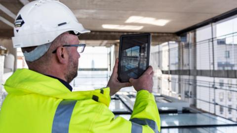 Architect in protective workwear on the building site is taking photos by his tablet.