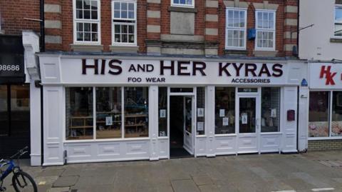Front of the old His and Hers footwear store, some of the letters from the sign are missing, the storefront is painted white.