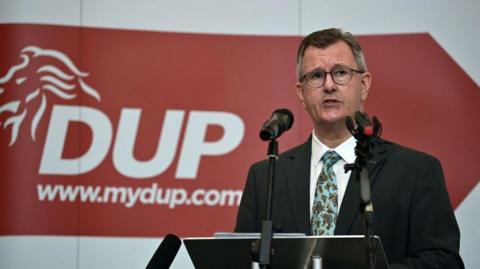 DUP leader Jeffrey Donaldson gives a keynote speech at the La Mon House Hotel on September 9, 2021 in Belfast, Northern Ireland. He is in front of two microphones wearing a dark suit, white shirt and patterned tie. A red sign is behind him with the words DUP www.mydup.com in white.