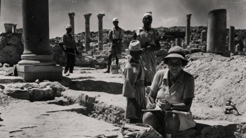 A black-and-white picture of a woman making notes while sitting in a trench in front of a number of ruined stone columns. A number of men stand behind her.