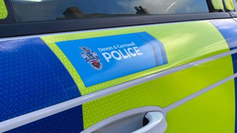 The side of a yellow and blue vehicle. There is a small blue sign on the side with the words 'Devon and Cornwall Police'. There is a window above the sign.