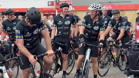 Image of a number of men in cycling gear, standing with their bicycles. In the background can be seen a crowd of people and the outside of a large stadium building