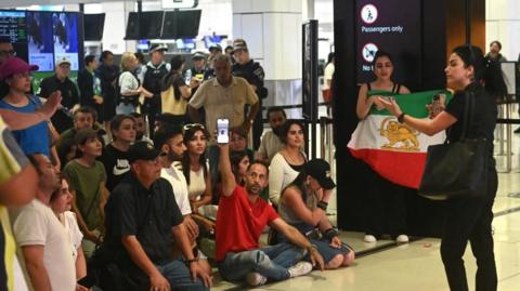 A group of around 15 people, some seated, some standing, at the airport. Some with the Lion and Sun flag, which served as the official state flag before the Islamic revolution in Iran