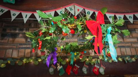Picture of a kissing bush hung from a ceiling in a home. It it wrapped with greenery and decorated with colourful ribbons and baubles. 