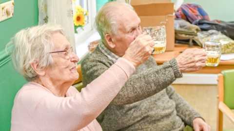 Two elderly people in a care home raising a glass of beer to cheers someone off camera