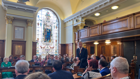 A man in a suit stands up speaking in an ornate council chamber full of elected members. There is a large stain glass window behind the councillors in Colchester City Council's main chamber.