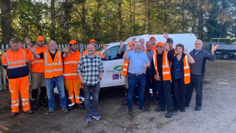 Sixteen people, mainly wearing orange workwear, smiling and waving towards the camera with trees, a white van and metal fencing in the background.