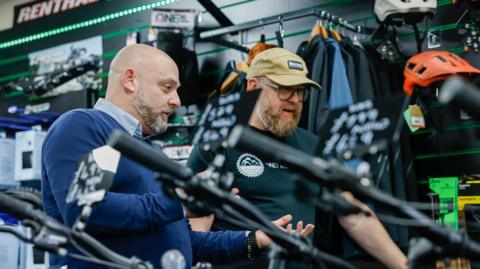 Paul Manuel advises a customer as they look at a range of cycles. Mr Manuel has a shaved head and short beard. He is wearing a blue jumper and light blue shirt. The customer, another man, has a beard and glasses, and is wearing a khaki baseball cap and black T-shirt. Accessories such as helmets can be seen hanging on the wall behind them.