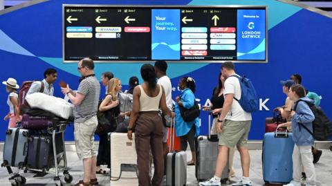 roughly a dozen people with luggage beneath information boards at Gatwick airport
