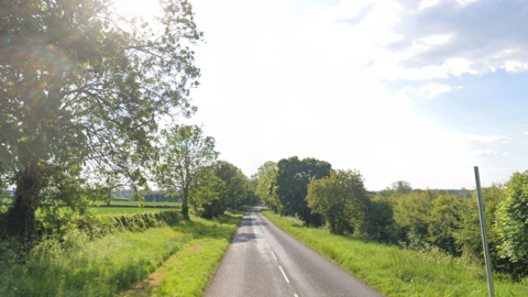 A straight road with a single lane on each side is pictured with grass and tress on each side on a cloudy but sunny day.