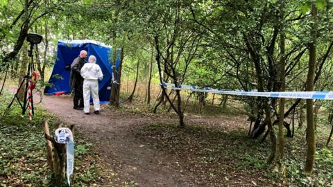 Police tape around a tree stump and stretching along several trees either side of a path. A forensic officer in a white suit and a police officer in a black coat are standing in front of a bright blue police tent to the left, with young green trees in the background