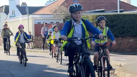 A group of children and adults cycle together along a residential street. The riders wear helmets and high‑visibility vests, and the scene appears to be part of an organised bike‑bus or school cycling activity on a sunny day.