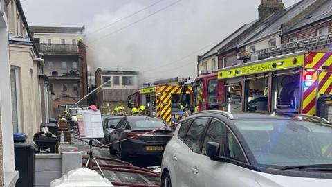 Two fire engines in a residential road with their blue lights on and firefighters getting equipment off the side. On one side of the road, cars are parked and smoke is billowing in the background