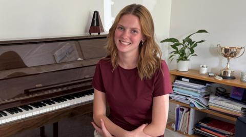 Freya Terris is a young woman with shoulder length light brown hair, which is hanging in loose waves around her face. She is smilling at the camera. She is wearing a burgundy t-shirt and is sitting on a piano stool with her arms crossed. She is sitting in front of a glossy brown piano, and there is a metronome, a plant and a large trophy seen sat around her. 