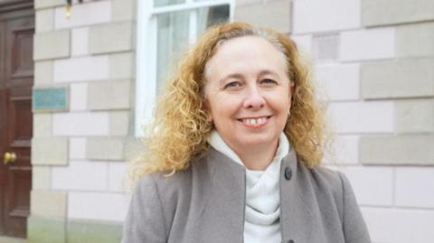 Jersey deputy Inna Gardiner has a broad smile while standing outside a government building. She is wearing a grey goat, white turtle neck jumper and has long curly blonde hair.