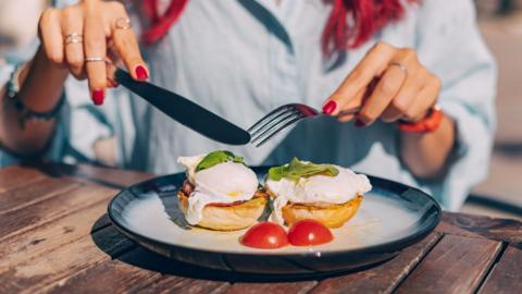 A person sits at a wooden bench eating poached eggs on a muffin. They have red painted and are wearing silver rings.