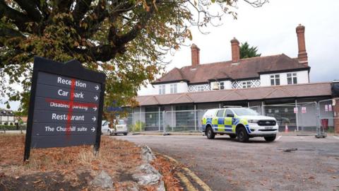 A hotel with a police SUV parked in front of it. The hotel which looks like a large house is surrounded by metal fencing. In the foreground a sign pointing to reception, parking and a restaurant has had a red cross painted on it so it resembles the st george's cross.