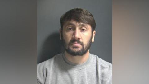 A police image of Max Pitt: a man with dark hair and a dark beard looking straight ahead and wearing a grey, round-necked t-shirt.