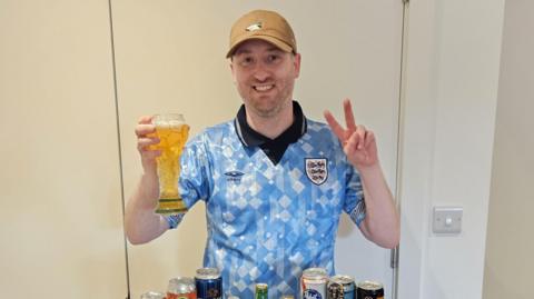 Gus Hully holds a beer in a glass shaped like the World Cup trophy, while he wears a vintage light blue England shirt and a brown baseball cap