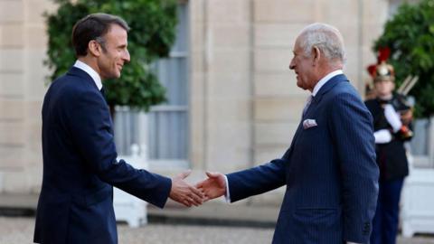 King Charles on the right of the picture holding out his right hand to President Macron, who is on the left of the picture, on the state visit to France in September 2023. Both men are wearing dark blue suits. 