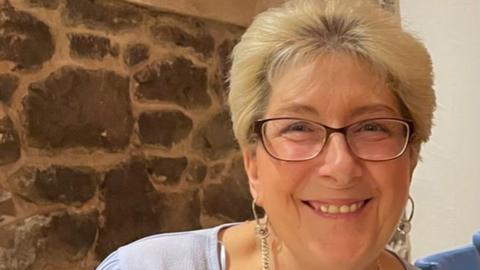 Sheena with short blonde hair wearing brown-rimmed glasses, a lilac top and dangly silver earrings standing in front of a stone wall smiling.