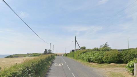 A rural road with trees and bushes down the side and a 30mph sign in the middle of the carriageway on a blue sunny day