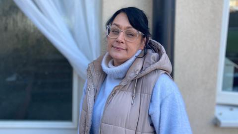 A woman with glasses, a beige gillet and grey rollneck jumper standing near a house