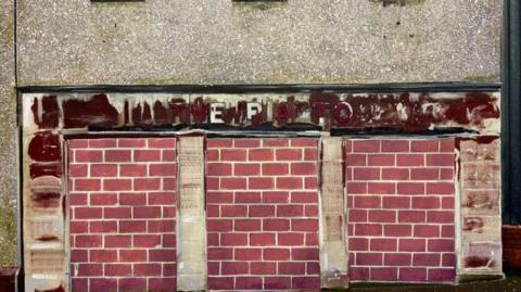 A painting by Narbi Price called "If you want to change things you cannit change them from the floor". It looks like a closed down business and there is an illegible sign painted above what look like bricked up doorways or windows.