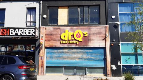 Dr Q's Food Factory which has a brown shop front with blue shutters which have not been pulled all the way down. A glass-fronted building can be seen to the right and a Turkish barber shop to the left. The flats would have sat above Dr Q's.