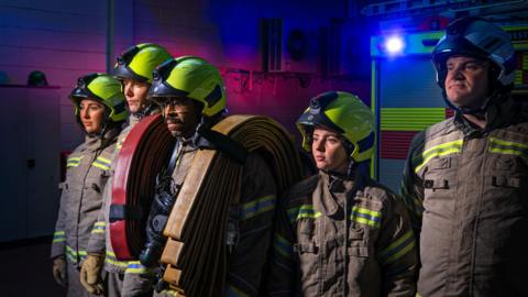 Five firefighters in brown fire suits with green reflective strips are standing in a line, in front of a fire engine. They are all wearing helmets. The firefighter in the middle has a roll of fire hose over each shoulder, and an oxygen mask around his neck