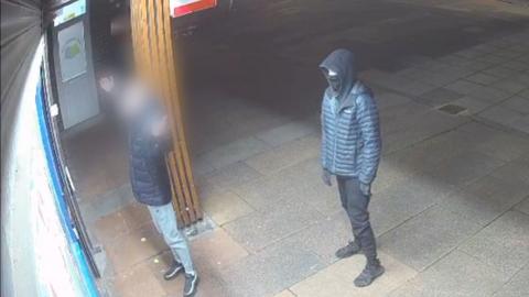 Two teenagers stand outside of a shop. One is wearing a face covering, the other's face has been blurred out. 