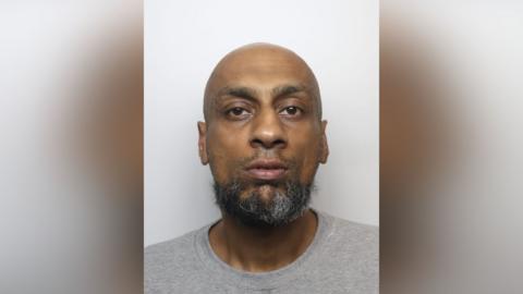 A bald man with a greying beard and grey t-shirt, pictured in a police mugshot. 