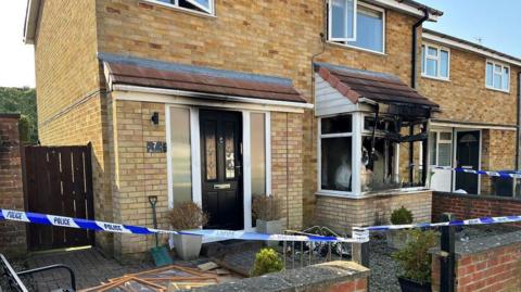 A two-storey, brick house which has suffered bad fire damage on the ground floor. The bay window has been smashed and inside it completely blackened. There is smoke damage above the front door.