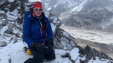 A woman in mountaineering clothes sits on the side of a mountain in Nepal, surrounded by snow. 