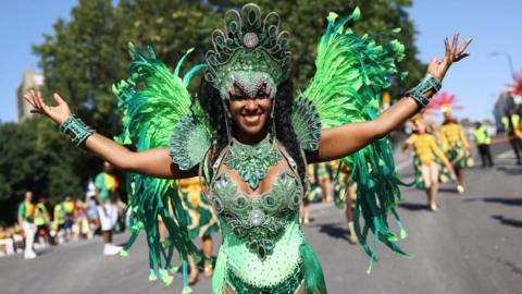 A woman stands in green sparkly carnival outfit with feathers and headdress, her arms are outstretched.