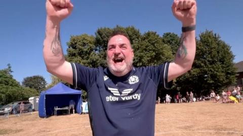 A man in a blue sports shirt raising his arms in the air in a field