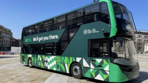 A green, black and white double decker bus parked in a square on a sunny day. 