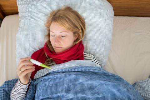 A young blonde woman is seen tucked up in bed holding a thermometer in a posed picture