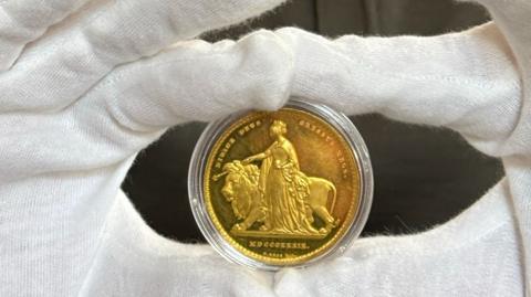 the coin in a clear case being held up between the index fingers and thumbs of a man wearing white gloves and a green shirt with his head down staring intently at the coin