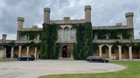 An exterior shot of Lincoln Crown Court which is a Gothic style building with turrets, greenery coming from it and large windows and a wooden front door. Two black cars are parked in front.