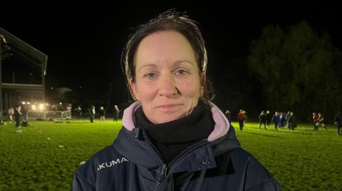 Jo pictured looking into the camera and smiling slightly. She's wearing a navy blue winter coat, with a black fleece on underneath. She's stood on a flood-lit rugby pitch, which has green grass, a dark night sky, and is bordered by trees. Behind her are about 15 girls running around and throwing rugby balls to one another. 