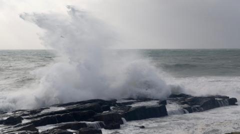 A tall wave crashes over rocks with an angry sea and grey sky in the background